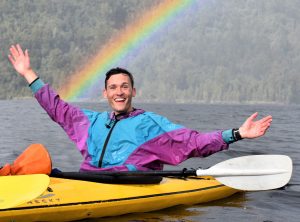 posing in a yellow kayak with a rainbow backdrop against the water and trees behind me