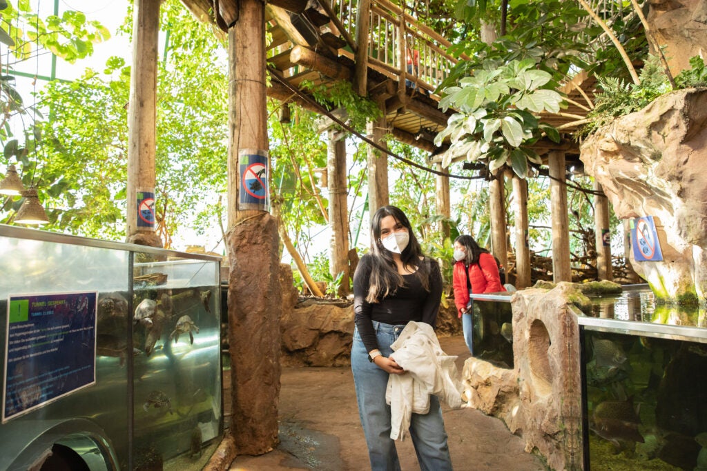 woman wearing face mask stands near trees, wooden pillars and tank with turtles