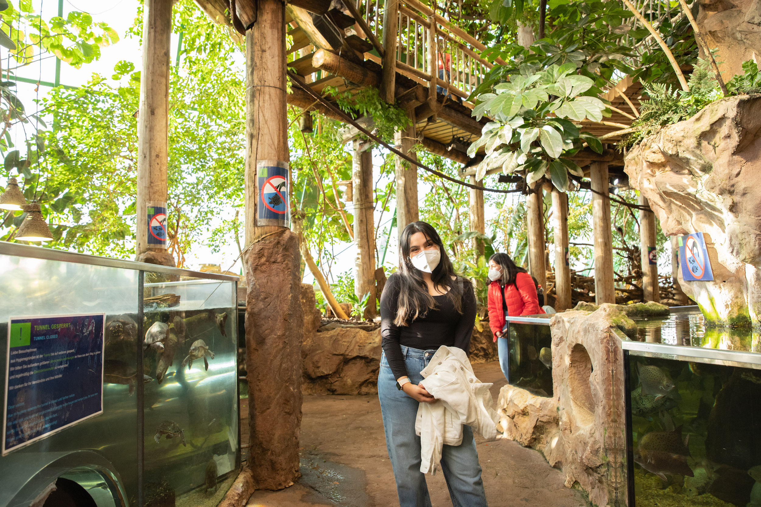 woman wearing face mask stands near trees, wooden pillars and tank with turtles 