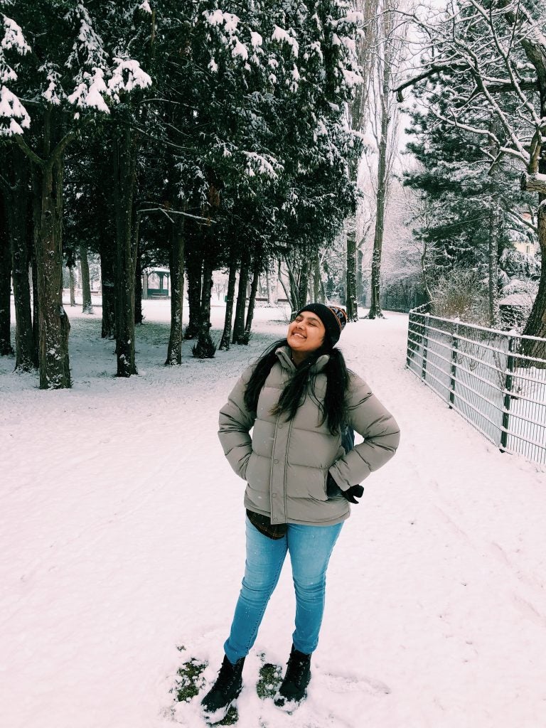 woman in winter clothes stands in snow looking up near trees during daytime