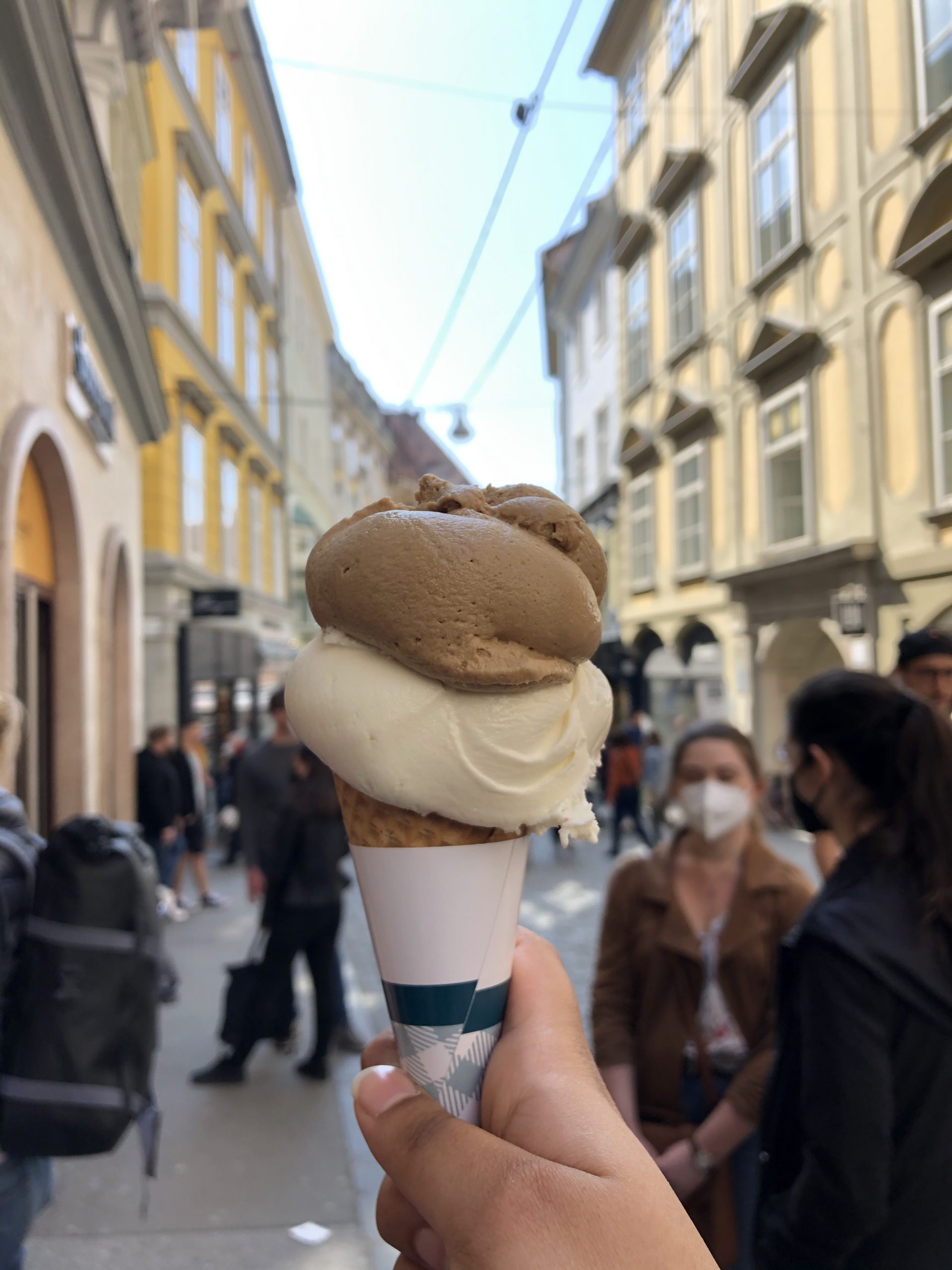 hand holding paper wrapped cone with gelato against backdrop of city street with people walking during daytime
