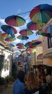 Rainbow umbrellas hang in the blue sky above a historic alleyway. 