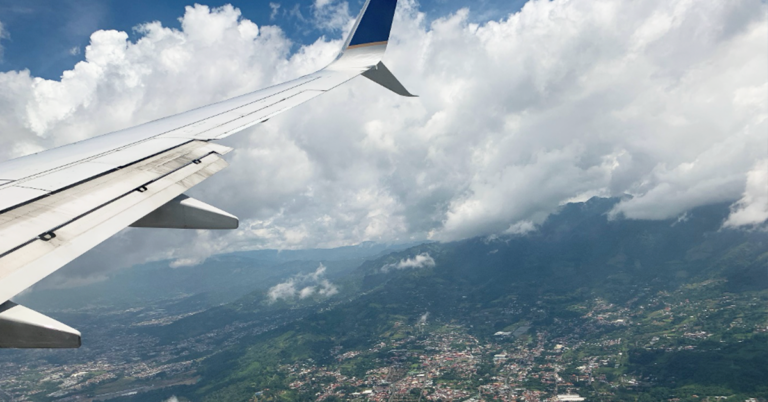 Birds-eye view from an airplane window of blue sky, white clouds and San Jose city below.