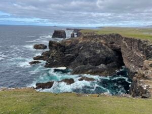 Waves crash into the rock and green grass of the Shetland Coast. 