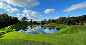 Bright blue sky with sparse white clouds is mirrored in a pond below, surrounded by manicured green lawn and forests.