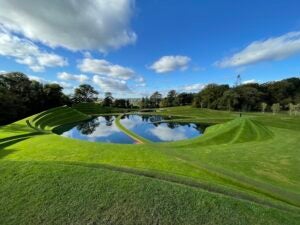 Bright blue sky with sparse white clouds is mirrored in a pond below, surrounded by manicured green lawn and forests. 