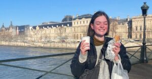 Eliza smiles in front of the the Pont des Arts in Paris, France. In one had she is holding a baguette and in the other, a capuccino