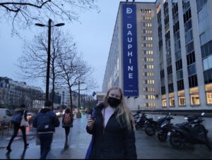 Kate stands in front of a building with a blue sign that reads "Université Paris Dauphine." She is wearing a black face mask.