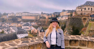Kate smiles in front of a view of the hills of Luxembourg with many buildings against a purplish sky. She is wearing a black hat and a hound's-tooth black and whiteblazer