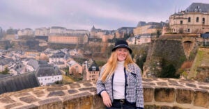 Kate smiles in front of a view of the hills of Luxembourg with many buildings against a purplish sky. She is wearing a black hat and a hound's-tooth black and whiteblazer