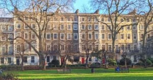 A student sits on a park bench in Gordon Square. The grass is bright green, but the trees behind the student are bare of leaves.