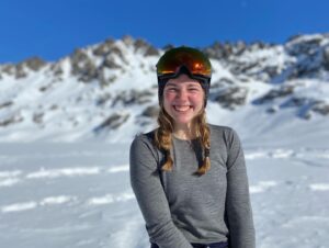 Katie smilesin front of a snowy alp in Switzerland wearing a gray long-sleeved shirt and snow hat