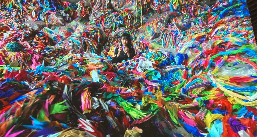 A screen grab from the origami paper crane video at the Mori Art Museum. It shows a Japanese woman in the middle of a pile of colorful origami cranes, which fill up the entire room