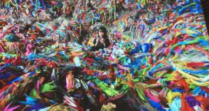 A screen grab from the origami paper crane video at the Mori Art Museum. It shows a Japanese woman in the middle of a pile of colorful origami cranes, which fill up the entire room