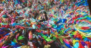 A screen grab from the origami paper crane video at the Mori Art Museum. It shows a Japanese woman in the middle of a pile of colorful origami cranes, which fill up the entire room