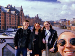 Jacki and four friends take a selfie near a body of water with a cityscape in the background