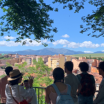 A group of students listen to a tour guide while gazing in front of them at the Spanish town and mountain range