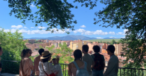 A group of students listen to a tour guide while gazing in front of them at the Spanish town and mountain range