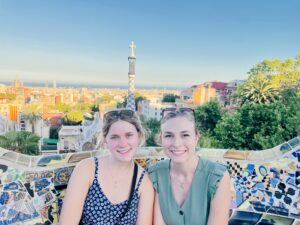 Darby and friend Halle sit together on a stone bench designed with brightly colored mosaic tiles, the buildings of Barcelona and the ocean behind them