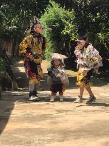 An adult and two kids dressed in elaborate fringe ensembles and with pig masks perform a traditional dance
