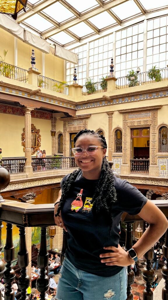 A girl with black shirt, standing in a brightly lit indoor space with ornate architectural details like railings, pillars, and tile work.