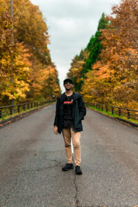Sourav smiles while standing in the middle of a street flanked by trees with vibrant, orange leaves