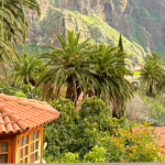 An outdoor landscape featuring a variety of palm trees, mountains, and steep slopes. On the side of the image is a small living structure with lots of windows and a red tiled roof.