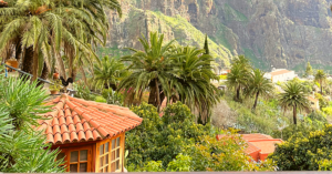 An outdoor landscape featuring a variety of palm trees, mountains, and steep slopes. On the side of the image is a small living structure with lots of windows and a red tiled roof.