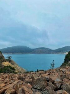 A calm, light blue lake with three mountains in the background and partly cloudy skies overhead.