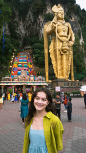 An girl stands in front of a Hindu temples and shrines, posing with a smile. She wears a blue dress while the entrance of the main cave features colorful details and many steps.