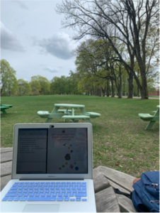 An open laptop placed on a wooden table in a park in Uppsala, Sweden with green trees and grass surrounding it.