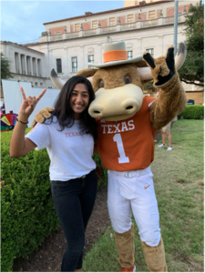 A student stands in front of a college building during the day, smiling and posing with the college mascot, who is wearing a burnt orange longhorn costume.
