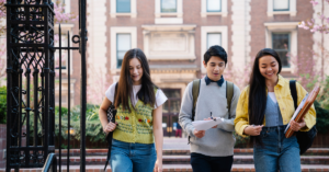 Three students walking together outside a school building on a sunny day, with backpacks on their shoulders and smiling faces, enjoying their time together.