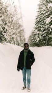 Man standing in a snowy landscape, wearing a black jacket, blue pants, and a green sweater with his arms by his side, excitedly grinning at the winter scenery.