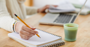 student using a pencil to write on a spiral notebook with a green coffee mug and laptop in the background.