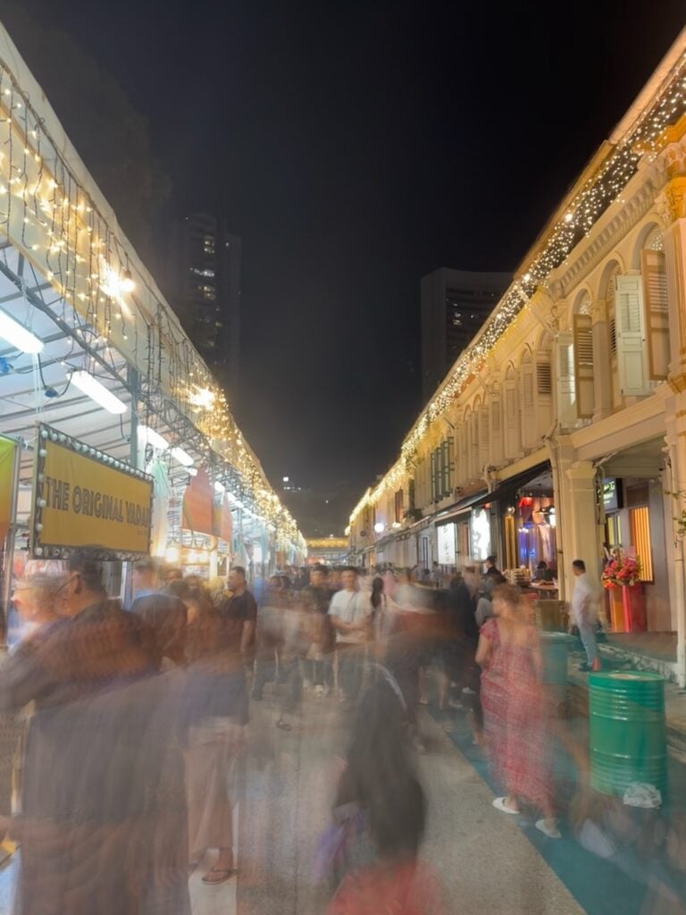 A night market with food stalls and people walking. The image is slightly blurred, adding to the lively and energetic atmosphere.