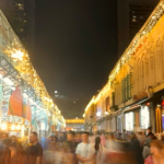 A night market with food stalls and people walking. The image is slightly blurred, adding to the lively and energetic atmosphere.