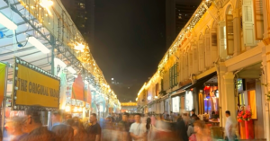A night market with food stalls and people walking. The image is slightly blurred, adding to the lively and energetic atmosphere.
