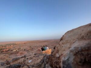 Four friends sit on a rock in the middle of a vast desert landscape and gaze out over a sprawling city in the distance.