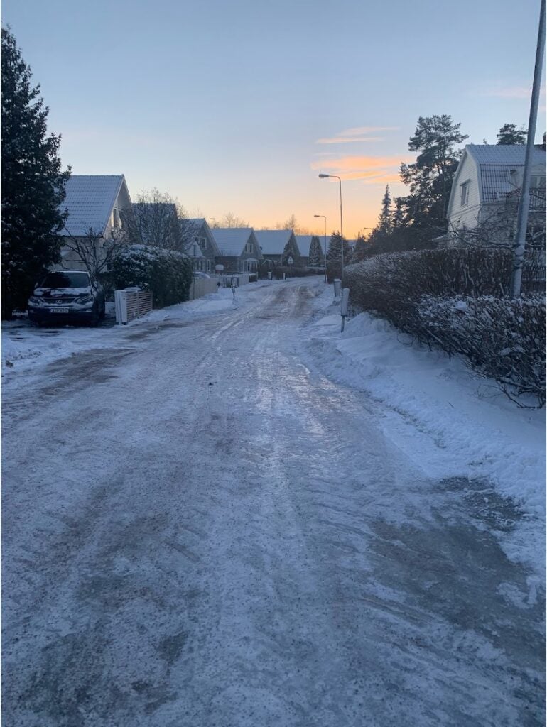 A winter scene of a neighborhood covered in snow with the sun peaking through clouds in the sky above.
