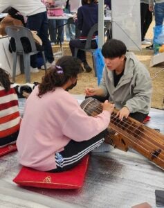 Two students sit cross-legged on the floor, facing each other, playing a traditional musical instrument.