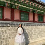 A student wearing a traditional South Korean dress, known as a hanbok, standing in front of a traditional building with intricate roof tiles and ornate wooden details on a sunny day.