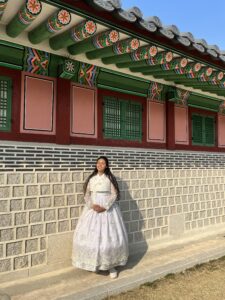A student wearing a traditional South Korean dress, known as a hanbok, standing in front of a traditional building with intricate roof tiles and ornate wooden details on a sunny day.