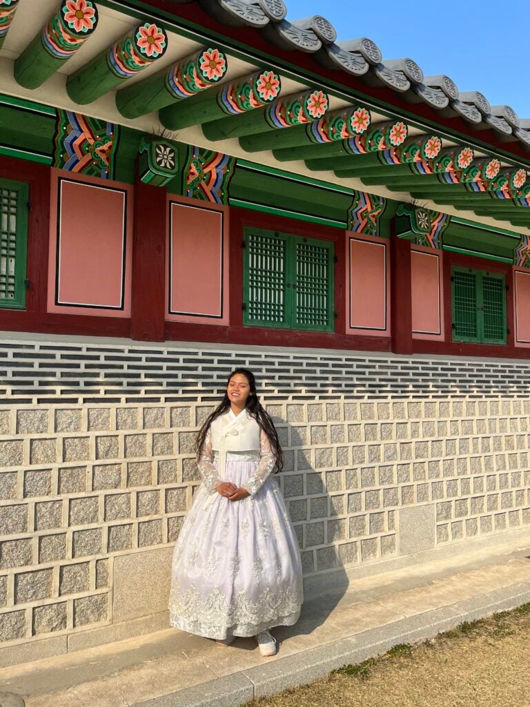 A student wearing a traditional South Korean dress, known as a hanbok, standing in front of a traditional building with intricate roof tiles and ornate wooden details on a sunny day.