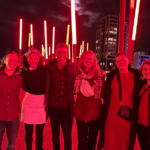 A group of smiling students posing on a bridge at night with vibrant red neon lights illuminating the background.