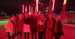 A group of smiling students posing on a bridge at night with vibrant red neon lights illuminating the background.