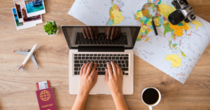 Aerial view of someone typing on a laptop computer with a map, cup of coffee, passport and photos surrounding the laptop.