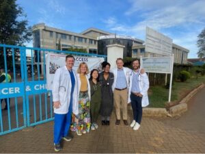 Doctors wearing white coats and scrubs pose with adults dresses in business professional attire in front of a hospital gate in Kenya.