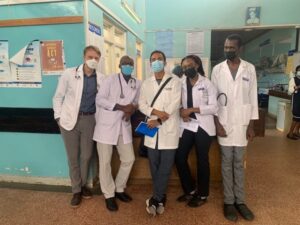 Five adults wearing white doctor coats wear face masks and pose inside a hospital.