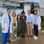 Doctors wearing white coats and scrubs pose with adults dresses in business professional attire in front of a hospital gate in Kenya.